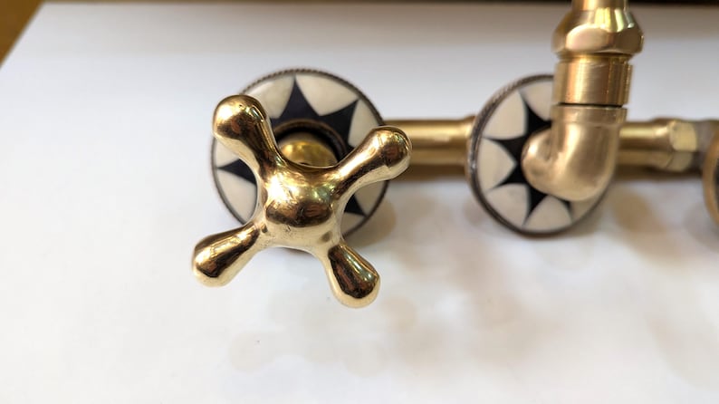 May include: Close-up of a brass faucet with a cross-shaped handle. The faucet has a gold finish and is set against a white background. The faucet's decorative elements include black and white starburst designs.