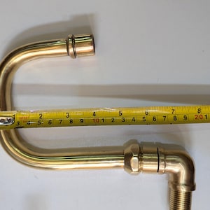 May include: A brass plumbing fixture with a curved design, approximately 10 inches long, is measured with a yellow tape measure. The fixture has threaded ends and appears to be new.