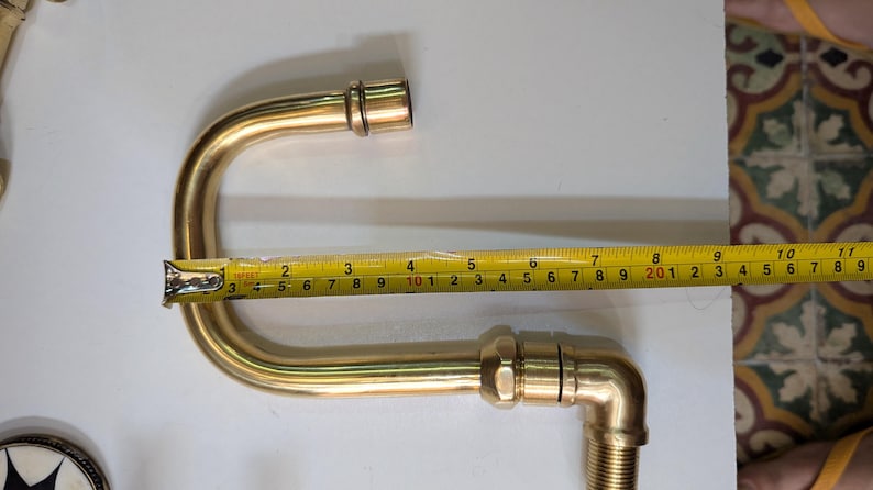 May include: A brass plumbing fixture with a curved design, approximately 10 inches long, is measured with a yellow tape measure. The fixture has threaded ends and appears to be new.
