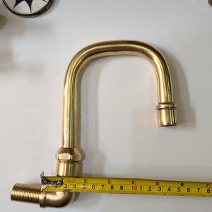 May include: A brass faucet with a curved spout, measuring approximately 7 inches in length, is shown with a measuring tape. Other brass fittings and a decorative circular object with a starburst design are also visible.