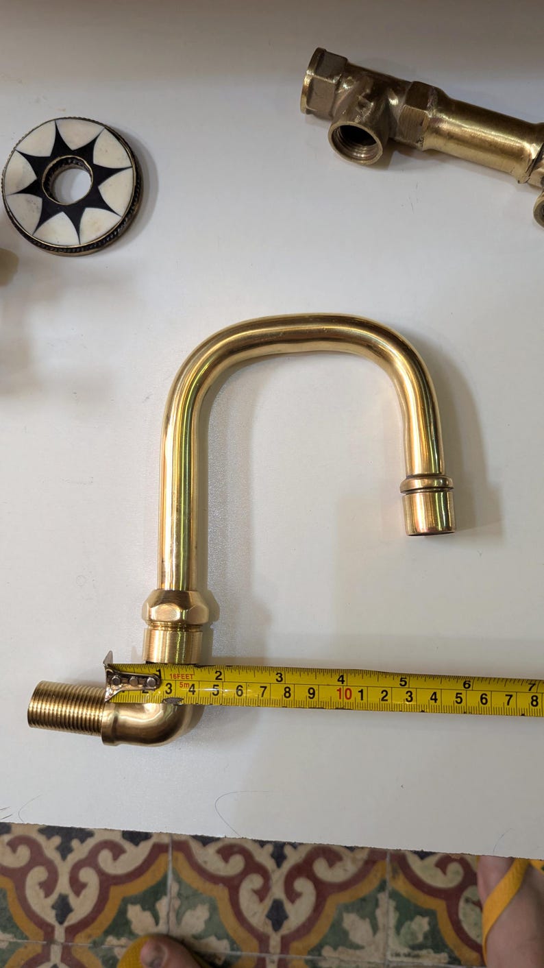 May include: A brass faucet with a curved spout, measuring approximately 7 inches in length, is shown with a measuring tape. Other brass fittings and a decorative circular object with a starburst design are also visible.