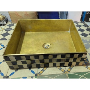 Könnte beinhalten: Ein rechteckiges Waschbecken mit goldener Innenausstattung und einer schwarz-weiß karierten Außenseite. Das Waschbecken hat einen Abfluss in der Mitte und ist aus Metall. Das Waschbecken steht auf einer gemusterten Fliesenoberfläche.