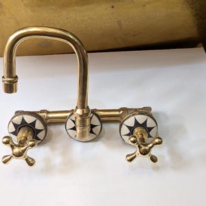 May include: A vintage-style brass faucet with a curved spout and three decorative handles. The handles feature a starburst design in black and white. The faucet is designed for a sink and has a classic, elegant appearance.