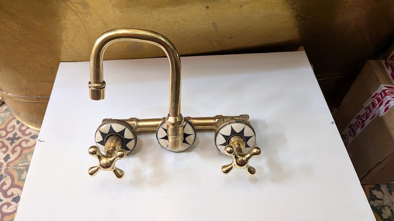 May include: A vintage-style brass faucet with a curved spout and three decorative handles. The handles feature a starburst design in black and white. The faucet is designed for a sink and has a classic, elegant appearance.