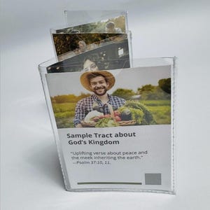 Può includere: Un espositore in plastica trasparente contenente opuscoli di esempio. L'opuscolo in alto presenta una persona sorridente con un cappello di paglia che tiene un cesto di verdure. Il testo sul fronte recita "Sample Tract about God's Kingdom" e un versetto biblico.