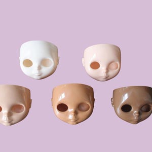 May include: Five doll heads in different skin tones, including white, light pink, tan, and brown, are displayed against a lilac background. Each head features large eye sockets and a small, detailed nose and mouth.