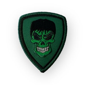 Puede incluir: Parche bordado verde en forma de escudo con una calavera verde con pelo negro y dientes blancos.