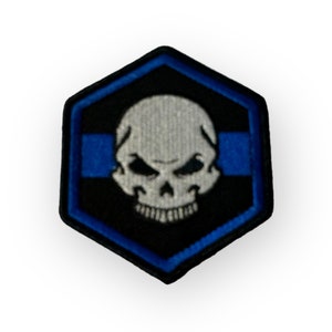 Op de afbeelding: Een zeshoekige patch in zwart en blauw met een wit schedel- en kruisbeenderontwerp. De schedel heeft een dreigende uitdrukking met een frons en scherpe tanden.