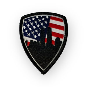 May include: A shield-shaped patch featuring an American flag design with a silhouette of a city skyline. The flag has blue, white, and red stripes and stars. The patch has a black border and a black city silhouette.