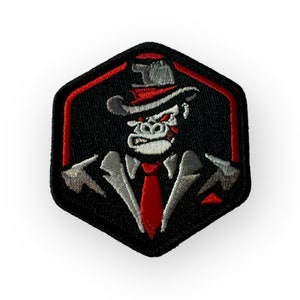 Pode incluir: Um remendo bordado preto e vermelho com um gorila de desenho animado usando um fedora cinza e uma gravata vermelha. O gorila tem uma expressão séria e está usando um terno cinza.