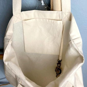 Op de afbeelding: Crèmekleurige canvas draagtas met een voorvak en lange banden. Een kleine bos sleutels hangt aan een bandje in de tas. Een doorzichtige glazen fles met een metalen dop is zichtbaar in de tas.