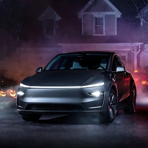 May include: A dark gray electric vehicle with a sleek, aerodynamic front profile, featuring a thin, horizontal LED light bar across the hood and bright, focused headlights. The car is positioned on a paved surface with a house in the background.