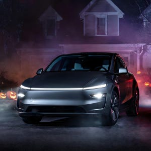 Könnte beinhalten: Ein dunkelgraues Elektrofahrzeug mit einem schlanken, aerodynamischen Frontprofil, das eine dünne, horizontale LED-Lichtleiste über der Motorhaube sowie helle, fokussierte Scheinwerfer aufweist. Das Auto steht auf einer gepflasterten Fläche vor einem Haus.
