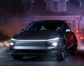 May include: A dark gray electric vehicle with a sleek, aerodynamic front profile, featuring a thin, horizontal LED light bar across the hood and bright, focused headlights. The car is positioned on a paved surface with a house in the background.