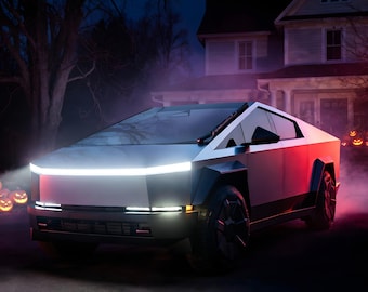 May include: A silver angular electric pickup truck with a prominent horizontal LED light bar across the front, dark wheels, and sharp geometric body panels, parked in a driveway at night with glowing jack-o'-lanterns in the background.