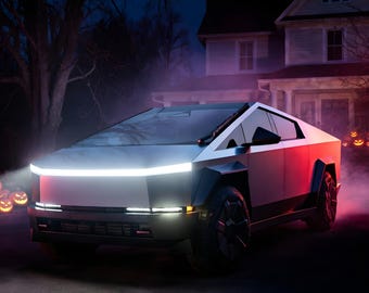 May include: A silver angular electric pickup truck with a prominent horizontal LED light bar across the front, dark wheels, and sharp geometric body panels, parked in a driveway at night with glowing jack-o'-lanterns in the background.
