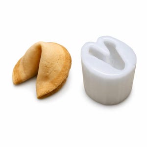 Puede incluir: Una galleta de la fortuna horneada junto a un molde de silicona blanco. La galleta de la fortuna es de color marrón dorado con una superficie texturizada. El molde es blanco liso, con forma de galleta de la fortuna.