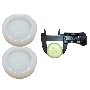 May include: Two white silicone molds for making round food items, such as cookies or cakes. A cucumber slice is being measured with a digital caliper, which reads 1.79 inches.