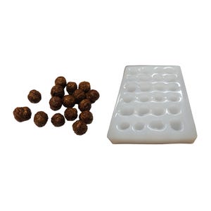 Peut inclure: Un moule en silicone blanc avec 24 cavités rondes, à côté d'un tas de petits objets ronds et bruns. Le moule est rectangulaire avec des bords arrondis. Les objets bruns semblent être de petits aliments sphériques.