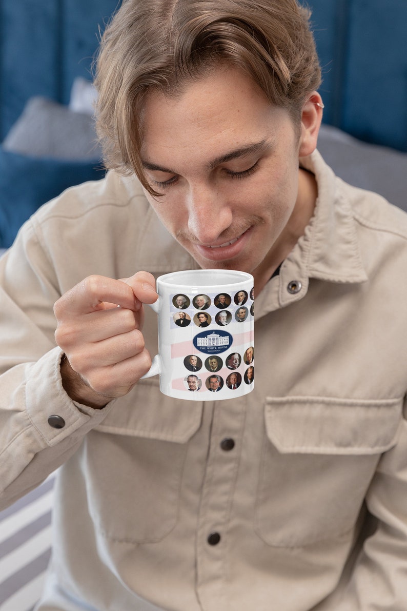 May include: White ceramic mug featuring portraits of U.S. presidents in circular frames. The mug has a design of the White House and the text "THE WHITE HOUSE WASHINGTON". The mug is held by a person wearing a beige shirt.