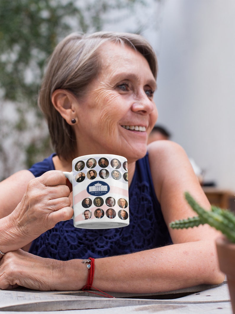 May include: White ceramic mug featuring portraits of U.S. presidents and the White House logo. The mug has a patriotic design with red and white stripes. The mug is held by a person.