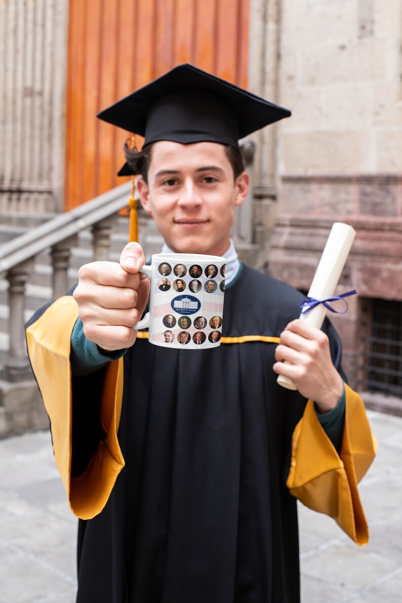 May include: A person in a graduation gown and cap holds a white mug featuring portraits of U.S. presidents and the White House logo. The person also holds a rolled diploma tied with a blue ribbon. The gown has gold accents.