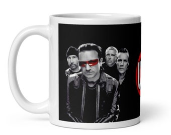 May include: White ceramic mug featuring a black and white image of a band with the lead singer wearing red-tinted glasses. The band is set against a black background. The mug has a white handle and a red and white logo on the side.