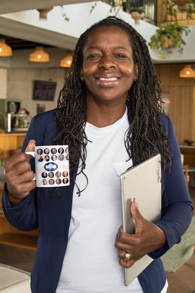 May include: White ceramic mug featuring portraits of U.S. presidents and the White House. The mug has a patriotic design with red and blue stripes. The mug is held by a person wearing a navy blue blazer and a white shirt.