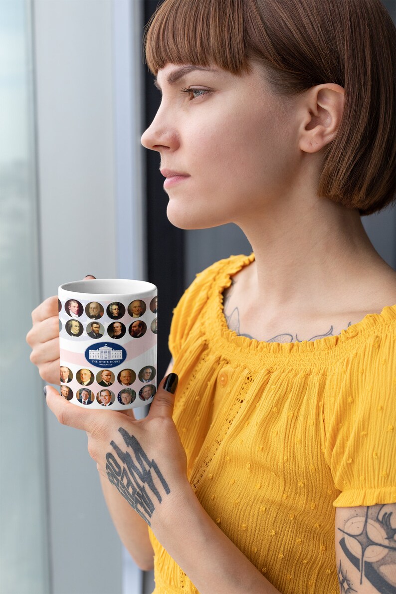 May include: White ceramic mug featuring portraits of U.S. presidents and the White House logo. The mug has a pink and white striped design. The mug is held by a person wearing a yellow top.
