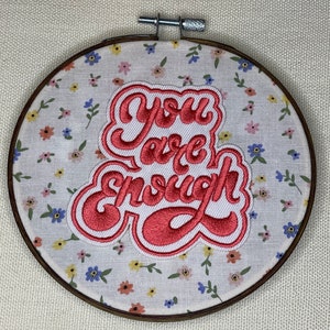 May include: A white embroidery hoop with a pink floral pattern. The hoop has a red embroidered patch with the text "You are enough" in a cursive font.