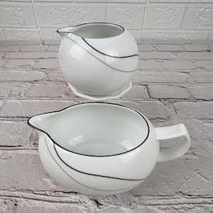 May include: Two white porcelain cream pitchers with black and gray accent lines. The top pitcher sits on a small, white saucer. The bottom pitcher has a handle. Both pitchers have a spout for easy pouring.