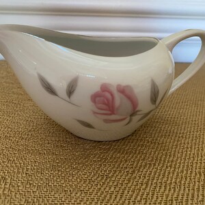 Puede incluir: Cremera de porcelana blanca con un delicado diseño de rosa rosa y hojas grises. La cremera tiene un pico curvo y un asa. Un borde dorado acentúa el borde. Mide aproximadamente 15 cm de largo.