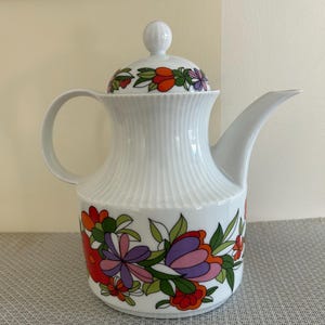 May include: White ceramic coffee pot with a lid and handle. The pot features a colorful floral design with red, orange, purple, and green flowers. The body of the pot has a ribbed texture.