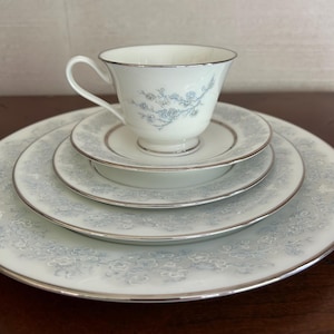 May include: A white porcelain teacup and saucer set with a delicate blue floral pattern and silver trim. The set includes a teacup, saucer, a small plate, and a larger dinner plate. The tableware is arranged on a wooden surface.