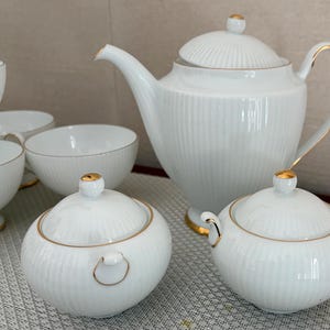 May include: A white porcelain tea set with gold trim. The set includes a teapot, sugar bowls, and teacups. The pieces have a ribbed design and are arranged on a light-colored surface.