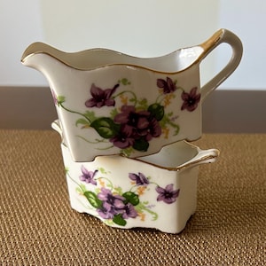 May include: Two white porcelain creamer pitchers with a floral design featuring purple and green flowers. The pitchers are stacked on top of each other.