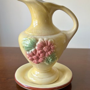 Könnte beinhalten: Ein kleiner, gelber Keramik Krug mit Blumenmuster. Der Krug hat einen Henkel und einen Sockel mit einer passenden Untertasse. Die Blumen sind rosa und grün.