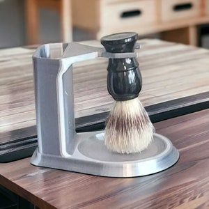 Puede incluir: Un soporte de plástico gris con un cepillo de afeitar negro y gris. El soporte tiene una ranura para el mango del cepillo y un pequeño cuenco para que el cepillo repose en él.