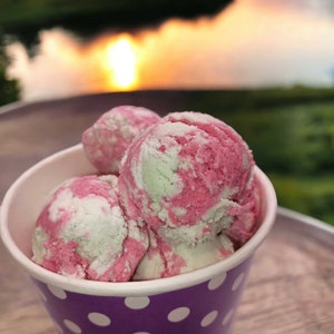 May include: A close-up of a purple and white polka dot paper cup filled with pink and white swirl ice cream. The ice cream is in a small cup and is sitting on a wooden surface.