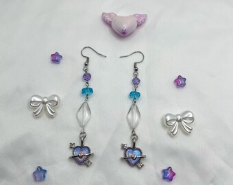 May include: A pair of dangle earrings featuring a heart charm, clear beads, and blue and purple glass beads. The heart charm has a galaxy design and is encircled by a silver ring.