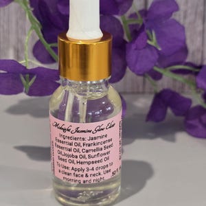 Puede incluir: Una botella de vidrio transparente de Midnight Jasmine Glow Elixir con tapa dorada y cuentagotas blanco. La etiqueta enumera ingredientes como Aceite Esencial de Jazmín e instrucciones de uso. El fondo presenta flores moradas.