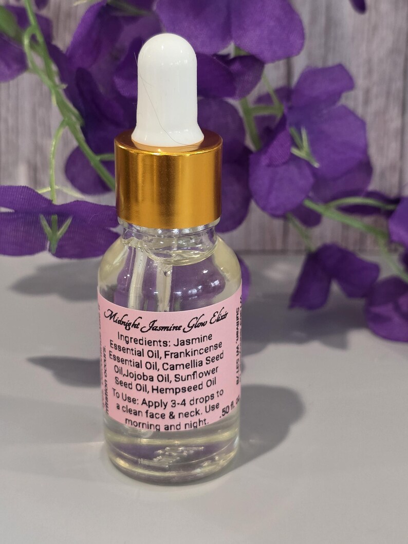 Puede incluir: Una botella de vidrio transparente de Midnight Jasmine Glow Elixir con tapa dorada y cuentagotas blanco. La etiqueta enumera ingredientes como Aceite Esencial de Jazm&iacute;n e instrucciones de uso. El fondo presenta flores moradas.