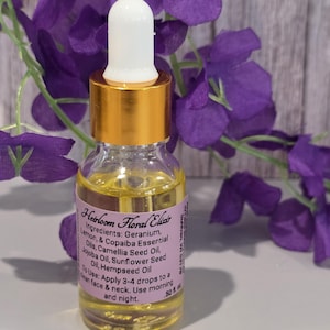 Puede incluir: Una pequeña botella de vidrio de Heirloom Floral Elixir con tapa dorada y cuentagotas blanco. La botella contiene un líquido amarillo y tiene una etiqueta con texto. El fondo presenta flores moradas.