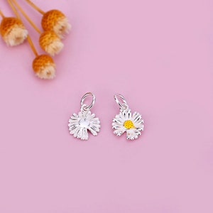 May include: Two silver daisy charms, one with a yellow center and one without. The charms are on a pink background.