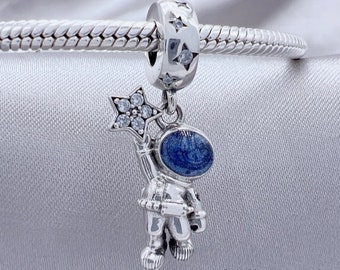 May include: Silver charm featuring an astronaut with a blue helmet and a star-shaped wand. The charm is suspended from a silver bracelet with a bead adorned with stars and clear crystals. The astronaut charm is detailed and whimsical.