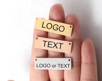 May include: Three rectangular metal tags in gold, peach, and silver, each with a small hole on either end. The top tag reads "LOGO" in black, the middle tag reads "TEXT", and the bottom tag reads "LOGO or TEXT".