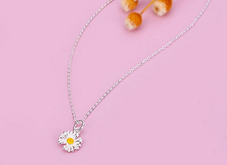 May include: A delicate silver chain necklace with a small white daisy pendant featuring a yellow center.
