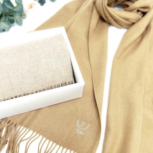 May include: A tan cashmere scarf with a white embroidered wreath and the name "Nan" on it. The scarf is folded and displayed in a white box with a tan scarf inside.