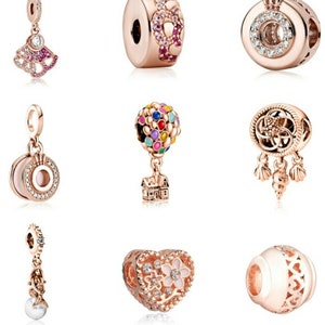May include: A collection of rose gold charm beads. Designs include a fan shape with pink gems, a bead with pink and purple gems, a round bead with clear gems, a charm with a hot air balloon and house, and a heart-shaped bead with a white flower.
