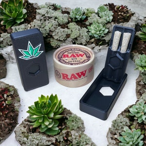 Puede incluir: Una caja rectangular negra con un diseño de hoja verde, un contenedor de cartón marrón con el texto "RAW Certified Authentic The Natural Way to Roll" y una caja rectangular negra con dos objetos cilíndricos blancos en su interior.